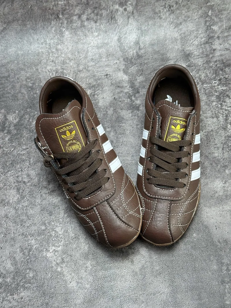 The Adidas Tokyo silhouette originates from the 1964 Tokyo Olympic lightweight track footwear line, positioned as a low-profile retro flat distinct from mass-market Samba OG and Gazelle. This full brown leather reissue launched late 2025, optimized for year-round lifestyle retail use. Compared to canvas vintage versions, grained cowhide improves durability and wear-in texture; the slim elongated last and thin gum sole create a slimmer foot silhouette without bulky padding.