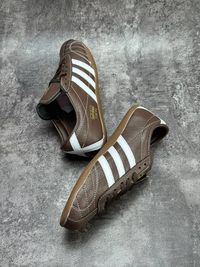 The Adidas Tokyo silhouette originates from the 1964 Tokyo Olympic lightweight track footwear line, positioned as a low-profile retro flat distinct from mass-market Samba OG and Gazelle. This full brown leather reissue launched late 2025, optimized for year-round lifestyle retail use. Compared to canvas vintage versions, grained cowhide improves durability and wear-in texture; the slim elongated last and thin gum sole create a slimmer foot silhouette without bulky padding.