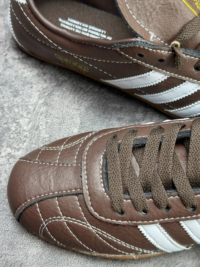 The Adidas Tokyo silhouette originates from the 1964 Tokyo Olympic lightweight track footwear line, positioned as a low-profile retro flat distinct from mass-market Samba OG and Gazelle. This full brown leather reissue launched late 2025, optimized for year-round lifestyle retail use. Compared to canvas vintage versions, grained cowhide improves durability and wear-in texture; the slim elongated last and thin gum sole create a slimmer foot silhouette without bulky padding.